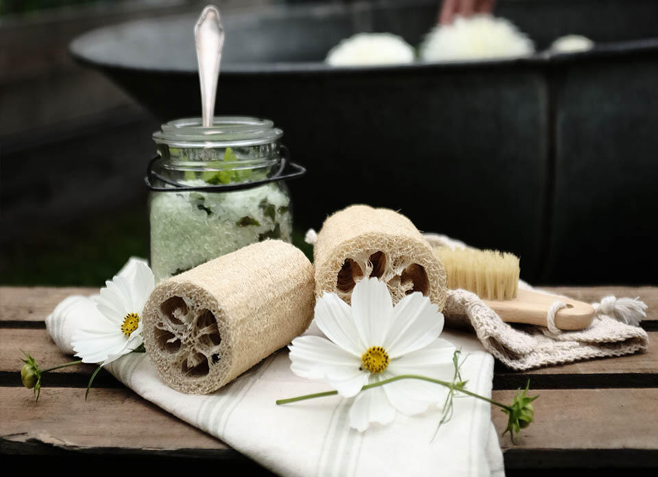 En person bak en krukke med hjemmelaget sukkerpeeling, blomster og en luffa.